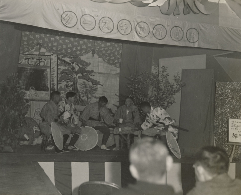 A black and white photograph of an indoor stage production. Men on the stage are dressed in traditional Japanese clothing.