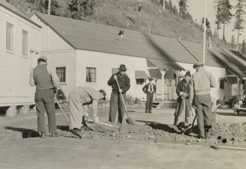 A black and white photograph of five men digging with shovels and pickaxes in front of buildings.
