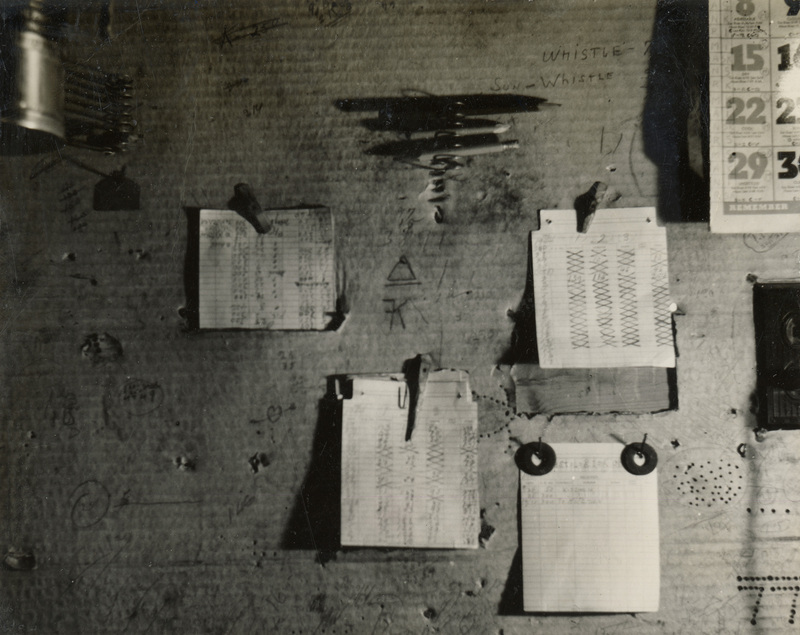 A black and white photograph of a billboard. There are four papers pinned to the board and a calendar at the top right.