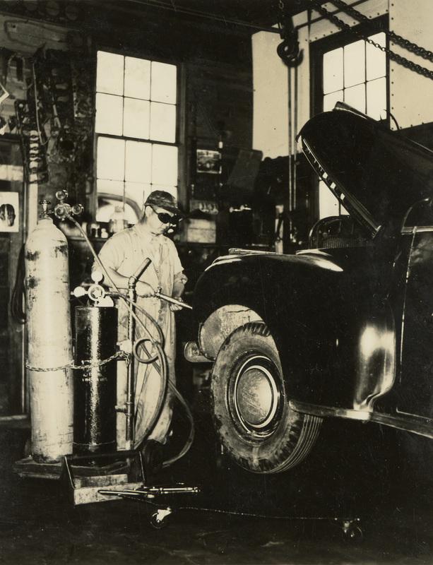 A black and white photograph of a man wearing protective goggles working on a car. The hood of the car is open and he is holding a tool toward the car.
