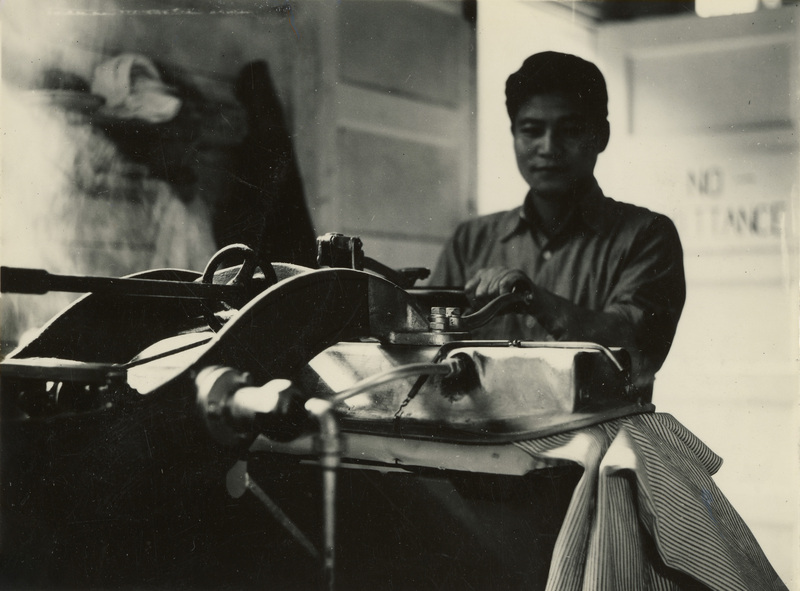 A black and white photograph of a man using a clothing press. The cloth in the press has a narrow striped pattern.
