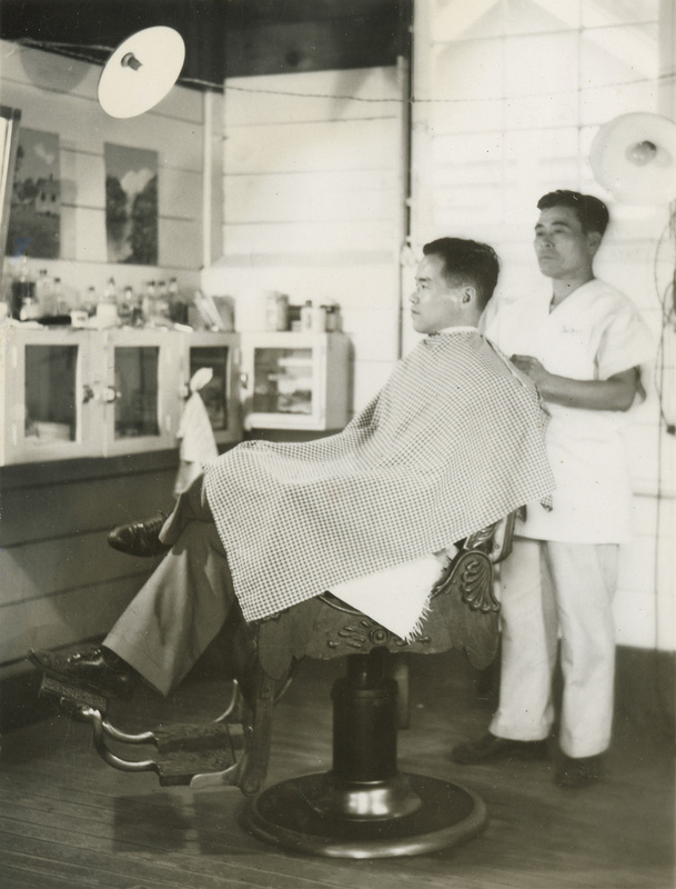 A black and white photograph of a man wearing a cape in a barber's chair. A male barber in all white stands behind the chair, to the right.