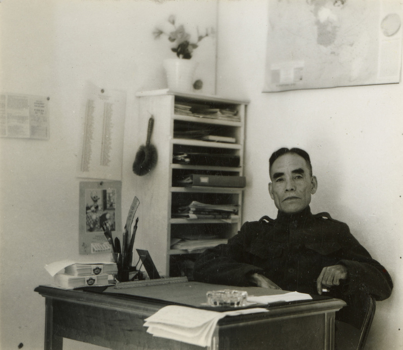 A black and white photograph of a man sitting at a desk. There is a bookshelf behind him and a map above him on the wall.