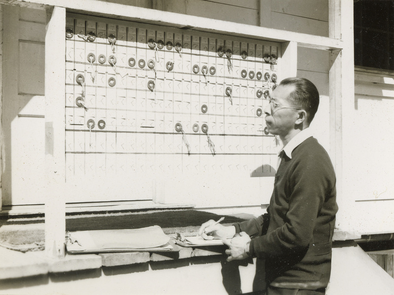 A black and white photograph of a man standing in front of a peg board used to keep track of work time.