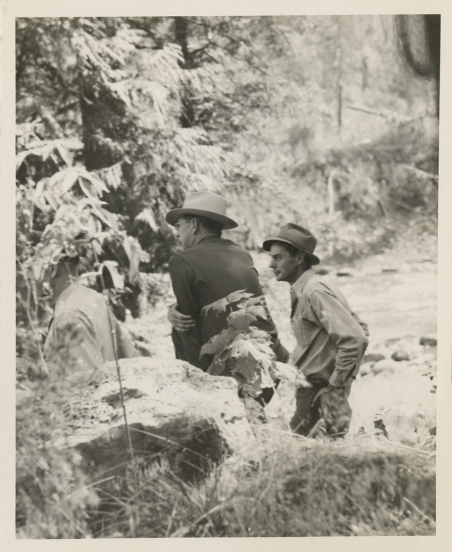A black and white image of three men standing by a river, talking to each other. There is a large rock in front.