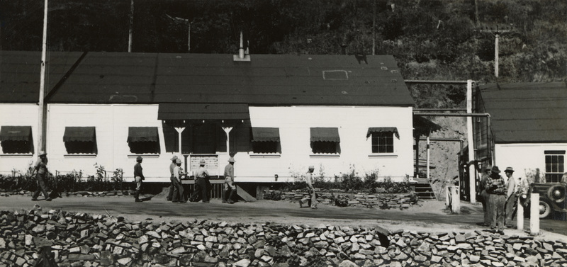 A black and white photograph of men walking in front of a long building.