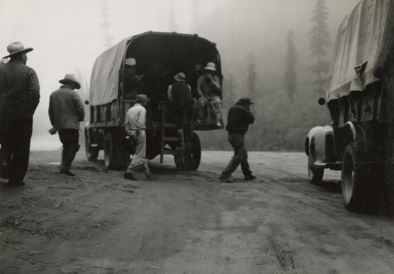 A black and white photograph of men standing behind a large transport truck. Men are also sitting inside of the truck.