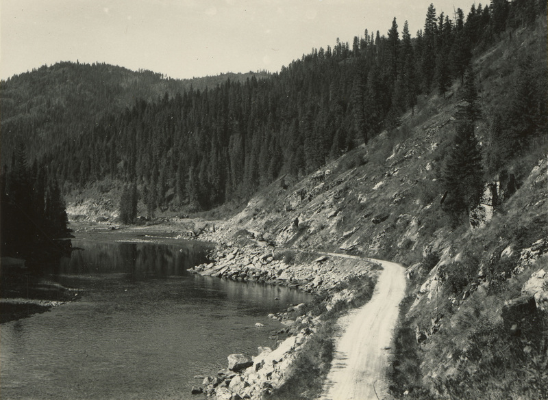 A black and white photograph of a dirt road running along a river.