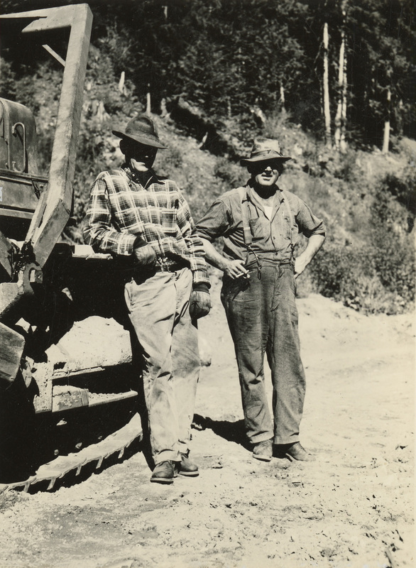 A black and white photograph of two men standing next to a bulldozer on a dirt road.