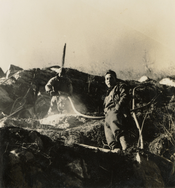 A black and white photogrpah of two men working on a hillside with construction tools, including a jackhammer.