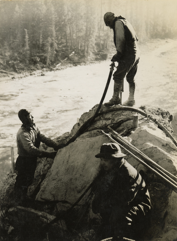 A black and white photograph of three men standing on a rock next to a river. They are holding hoses and construction equipment.