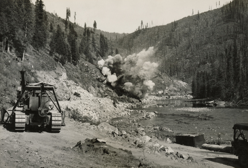 A black and white photograph of an explosion along a river, with construction images along the road where the explosion is occurring.