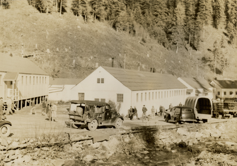A black and white photograph of vehicles in front of buildings next to the river.