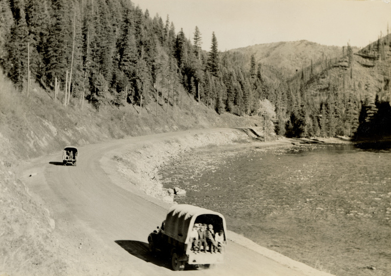 A black and white photograph of two vehicles driving on the dirt road along a river.