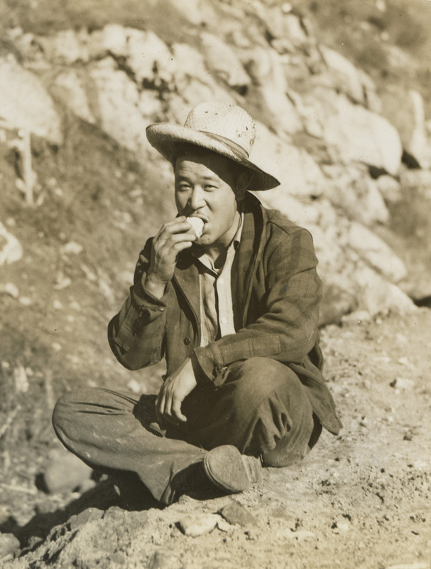 A black and white photograph of a man sitting with crossed legs and eating lunch.