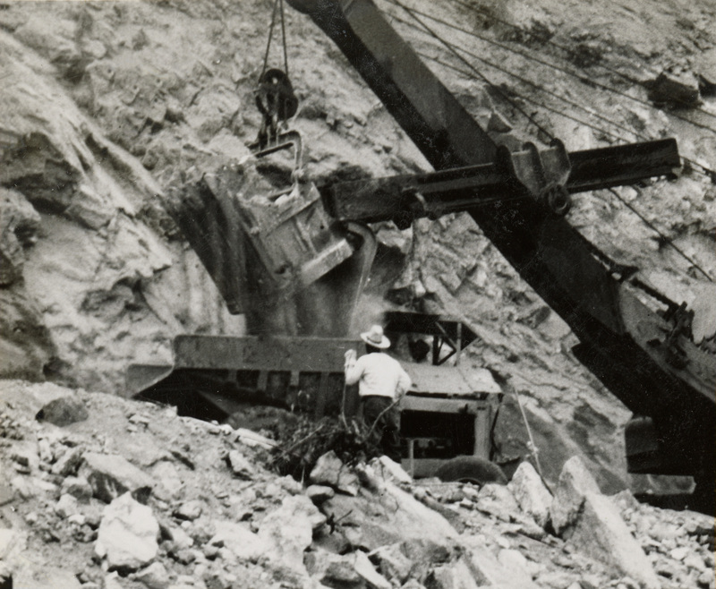 A black and white photograph of a crane moving dirt on a hillside. A man in a white shirt stands in front of the crane.