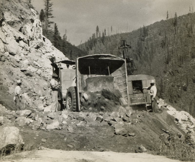 A black and white photograph of construction equipment full of dirt on a hillside. There are men standing in front of the equipment monitoring the process.