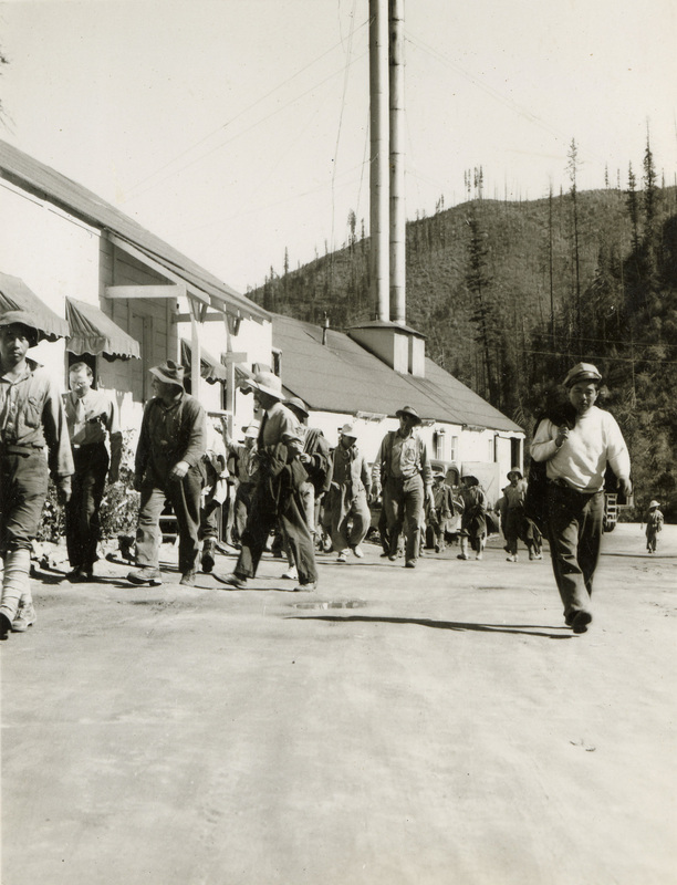 A black and white photograph of many men walking while looking at the side of a building. Most of the men are wearing helmets or hats.