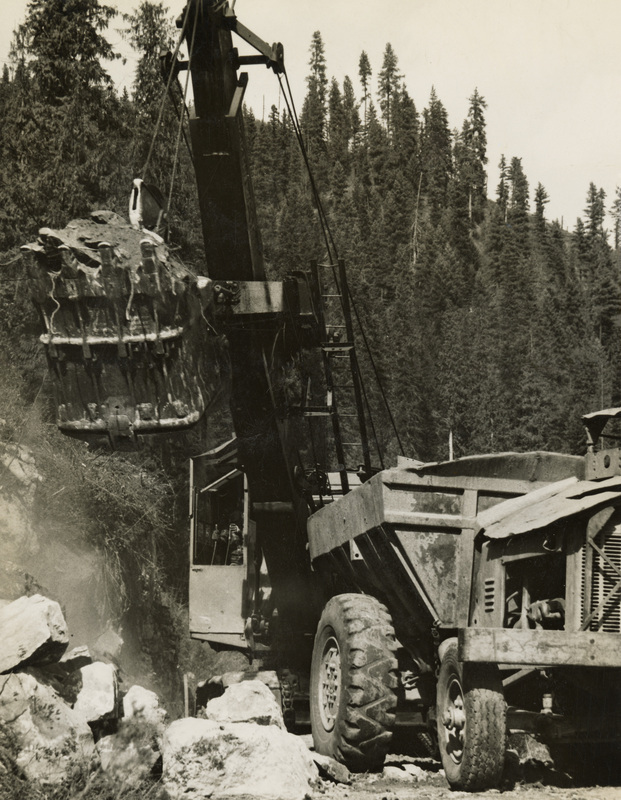 A black and white photograph of a crane and other construction equipment moving large rocks and dirt from the road.