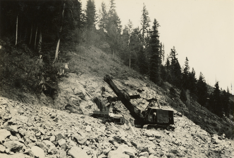 A black and white photograph of a crane on a rocky hillside. The tree line is above the crane itself.