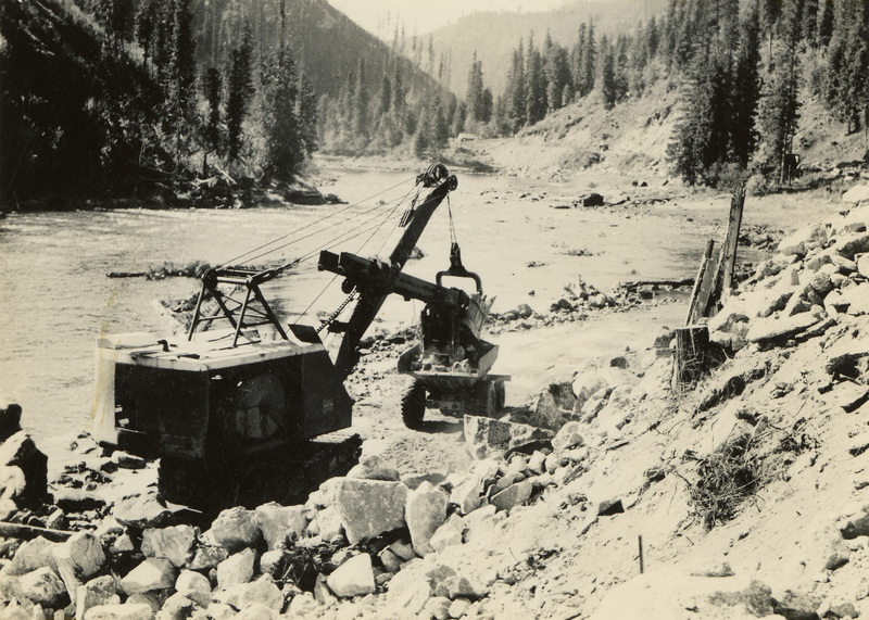 A black and white photograph of a crane with a large log in its grasp. The crane is next to a scenic view of a river.