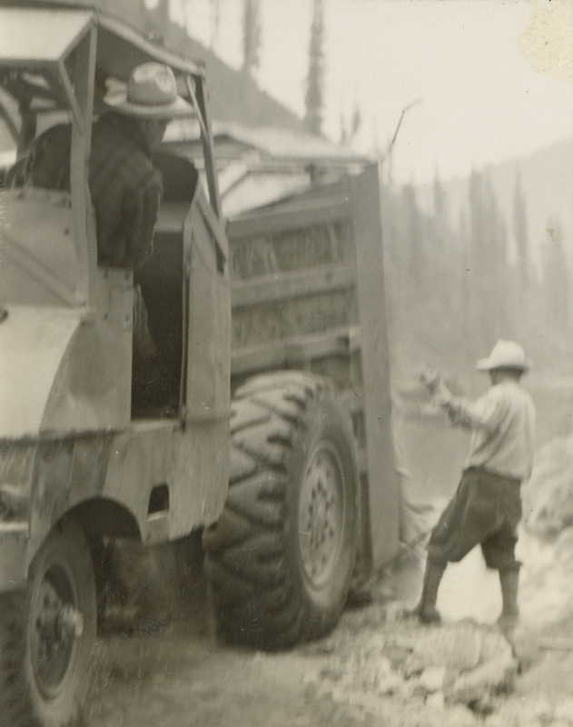 A black and white photograph of a dump truck. A man stands toward the back of the truck and is directing a man in the driver's seat of the truck.