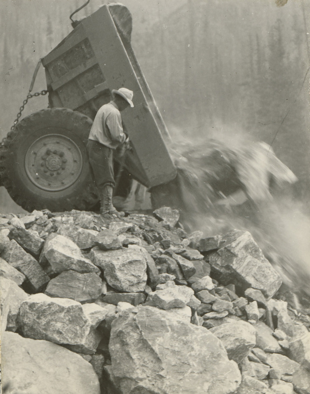 A black and white photograph of a man unloading a dump truck full of rocks.