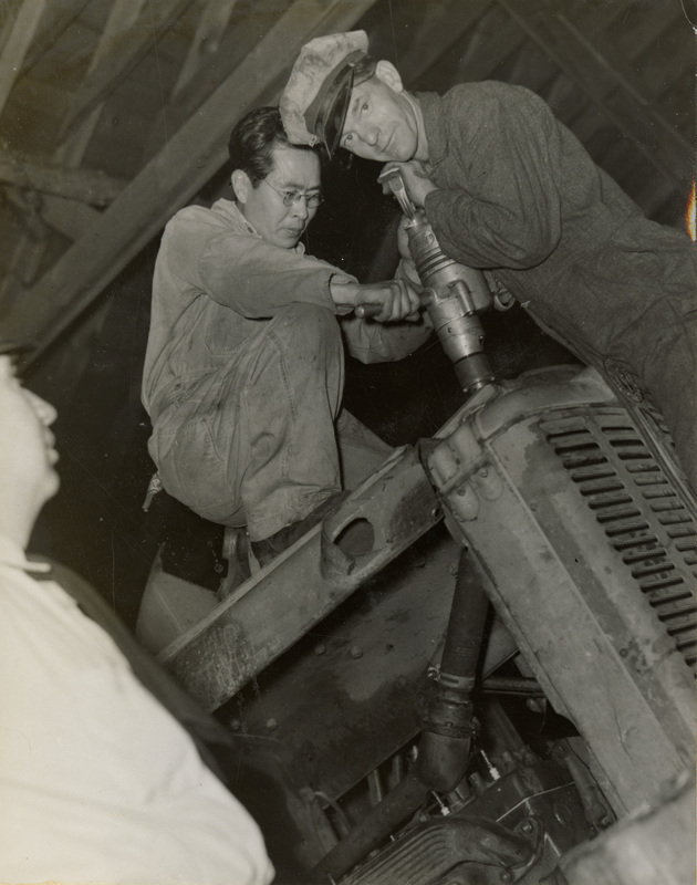 A black and white phtoograph of three men working on a tractor or another piece of machinery.
