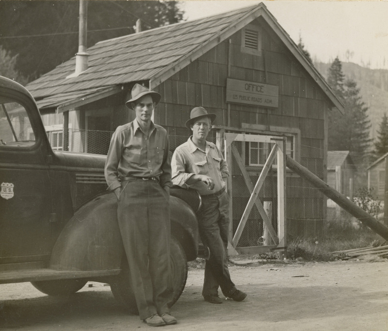A black and white photograph of two men leaning against a vintage truck in front of a wood building.