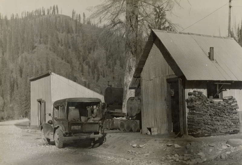 A black and white photograph of a vehicle parked in front of two buildings. The closest building on the right has a large stack of firewood.