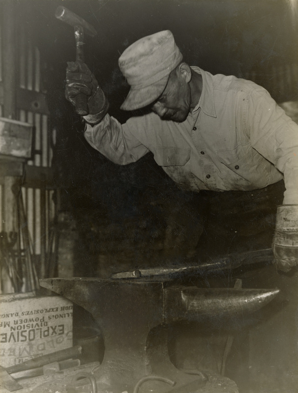 A black and white photograph of a man who is about to hit an anvil with a hammer.