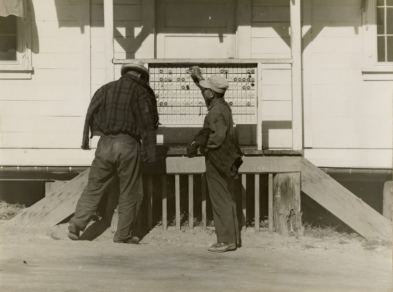A black and white photograph of two men outside a building looking at a peg-board used for checking in for work.