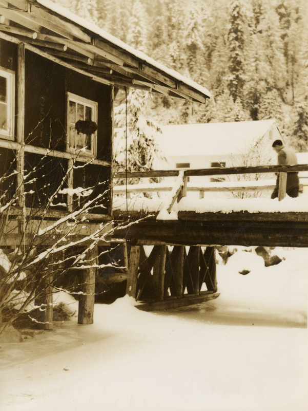 A black and white photograph of a man crossing a snow-covered bridge to a log building.