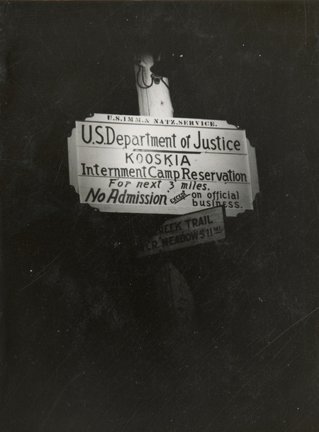 A black and white image of a sign. It reads "US Department of Justice. Kooskia. Internment Camp Reservation. For next 3 miles. No Admission except on official business." A smaller sign sits below it but is too dark to read.