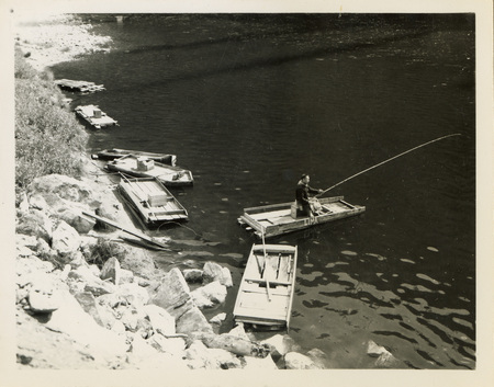 A black and white image of a man fishing near a river. Two boats sit along the bank at the river.