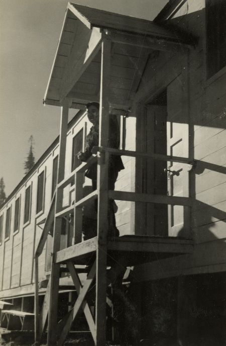 A black and white photograph of a man standing on a high porch of a building. He is leaning along the railin gof the porch.