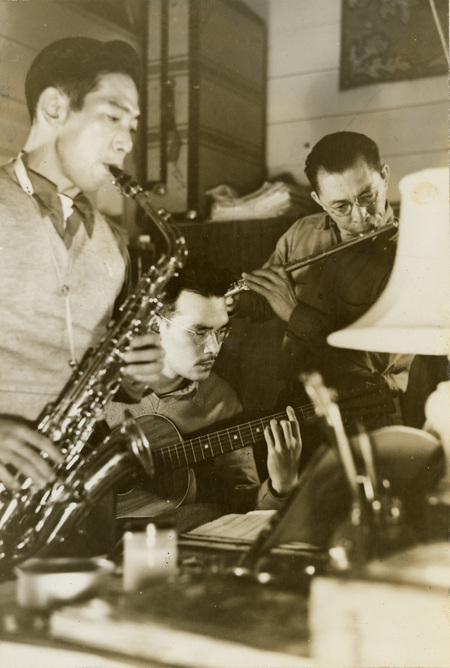 A black and white photograph of three men playing instruments: one playing a saxophone, one playing a guitar, and one playing a flute. 