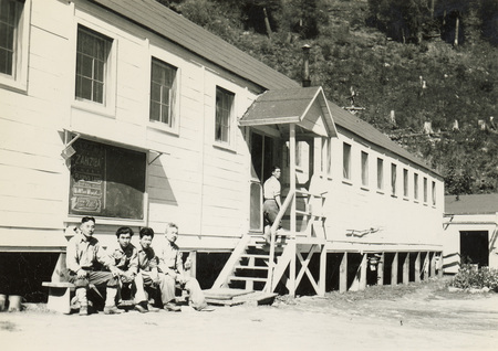 A black and white photograph of a group of men sitting on a bench in front of a building. Another man is pictured walking up a set of stairs to the entrance of the building.