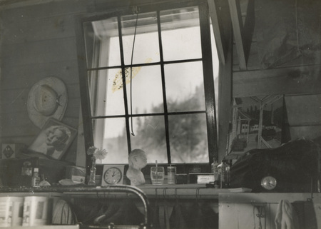 A black and white photograph from the inside of a window. A number of items such as a bust of a woman, glasses, posters, and a clock sit below the window.