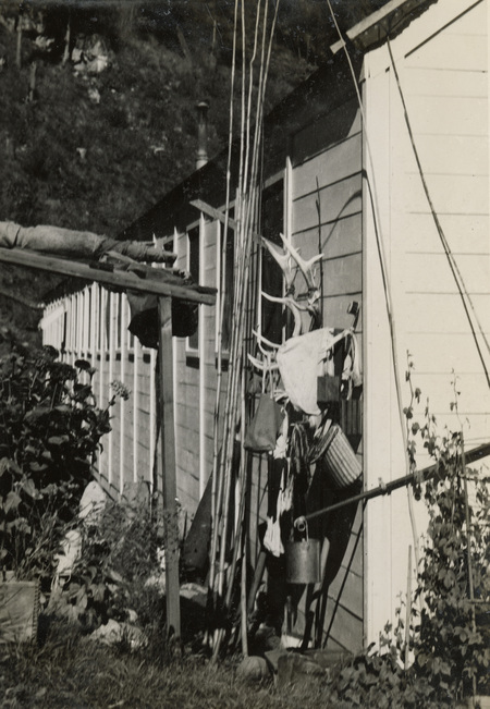 A black and white photograph of the outside of a building with fishing and hunting items leaning against the siding of the building. There are plants, grass, and shrubs along the building.