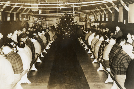 A black and white photograph of men sitting along two long tables on either side. They are eating dinner.