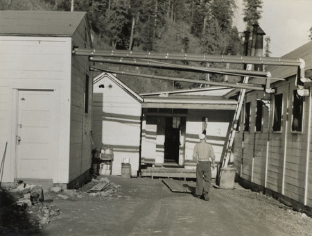 A black and white photograph of the outside of buildings at the Kooskia Internment Camp. A man is walking down an alley.