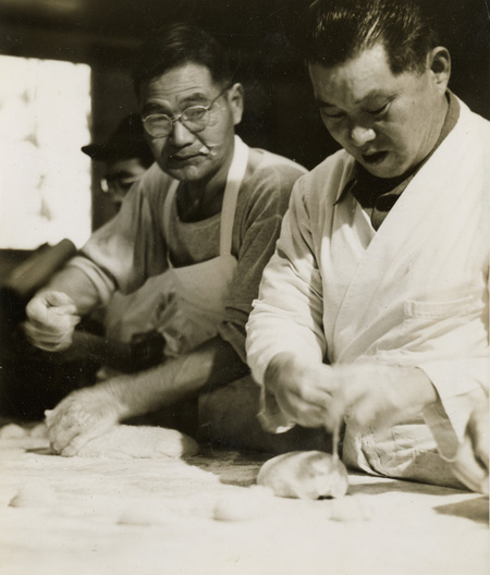 A black and white photograph of two men preparing food. They appear to be making bread.