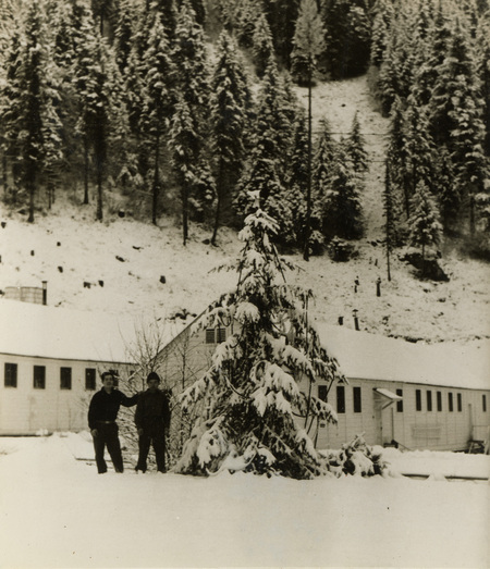 A black and white photograph of two men standing outside in the snow. There is a pine tree in front of the buildings that they are standing next to.