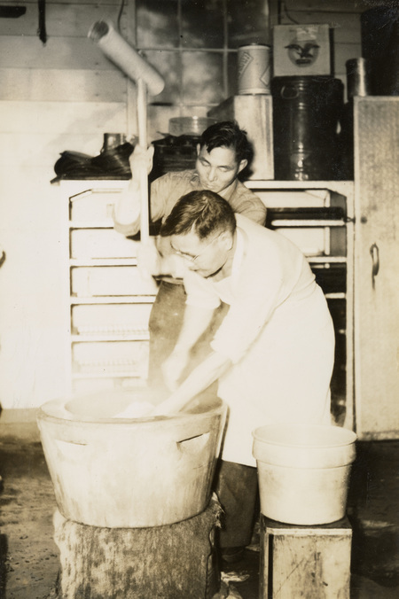 A black and white photograph of two men making mochi in an usu.
