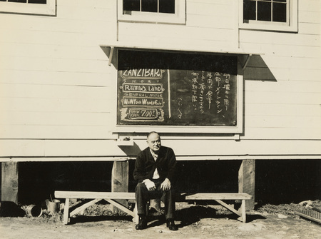 A black and white photograph of a man sitting under a chalkboard on the outside of a building. Both English and Japanese can be read from the sign.
