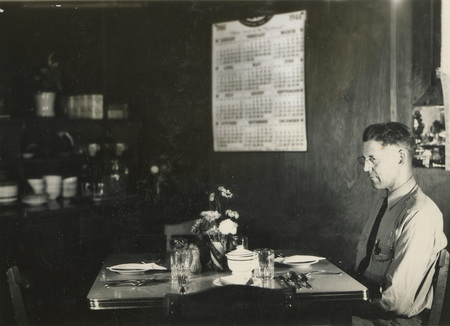 A black and white photograph of a man sitting at a table with dinnerware in front of him. A large calendar of the twelve months hangs on the wall.