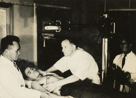 A black and white photograph of a patient on a bed, with three medical personnel attending to the patient on the table. 
