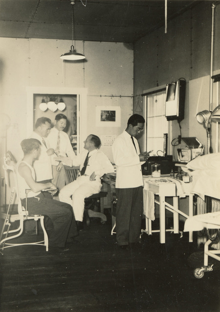A black and white photograph of five men in a medical office. Two of them are sitting on chairs.
