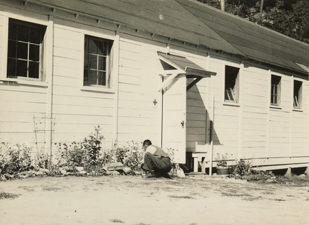 A black and white photo of a man kneeling in front of a flower bed at the base of a building.